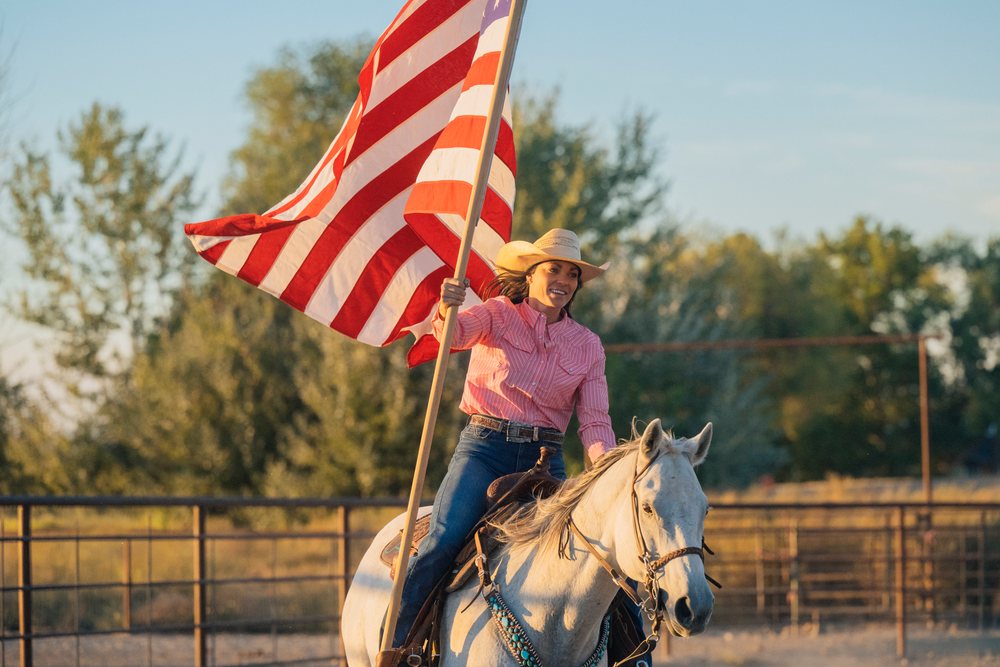 Woman riding a white horse while holding an American flag in an outdoor riding arena in the Southwest USA at sunset.