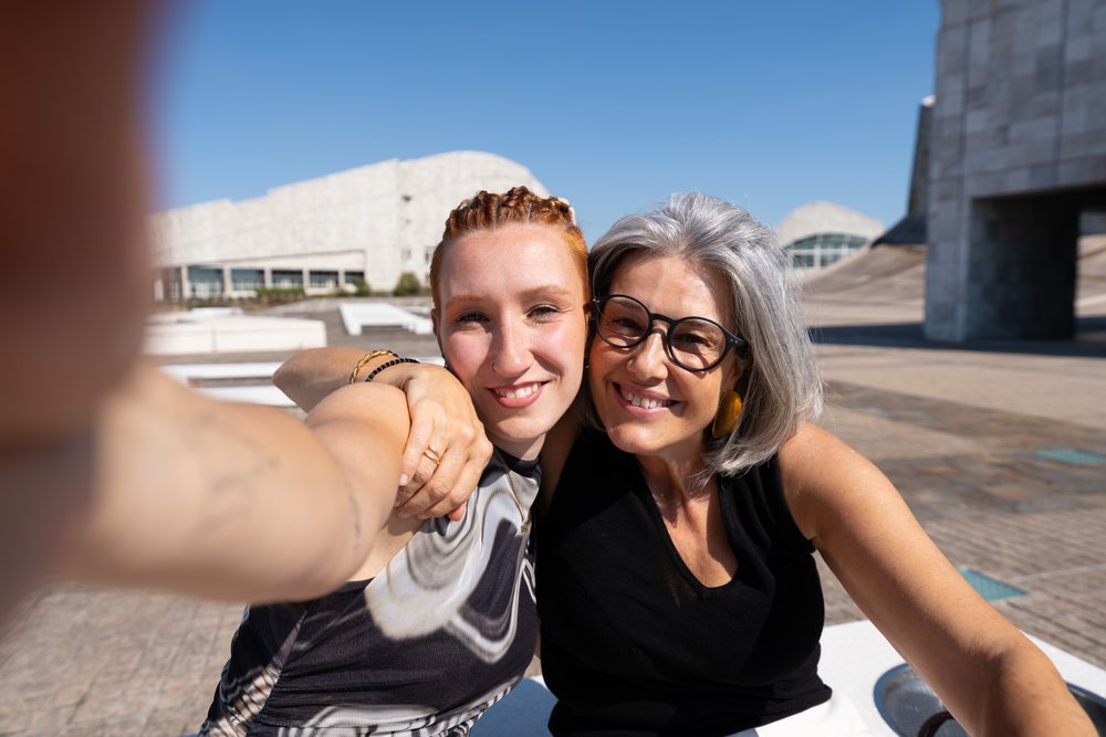 Two women smiling and embracing while taking a selfie outdoors in an urban setting, businesswomen, Spain.