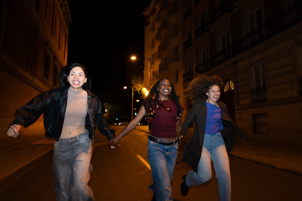 Three smiling women holding hands and running joyfully on a paved urban street in Madrid at night, Spain.