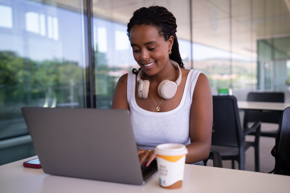 A woman with headphones around her neck types on a laptop at a desk with a coffee cup in a modern office workspace, remote work, travel, Santiago De Compostela, Spain.