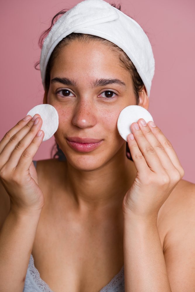 Woman gently cleanses her face with cotton pads against a plain pink background wearing a white headband, beauty shoot, Spain.