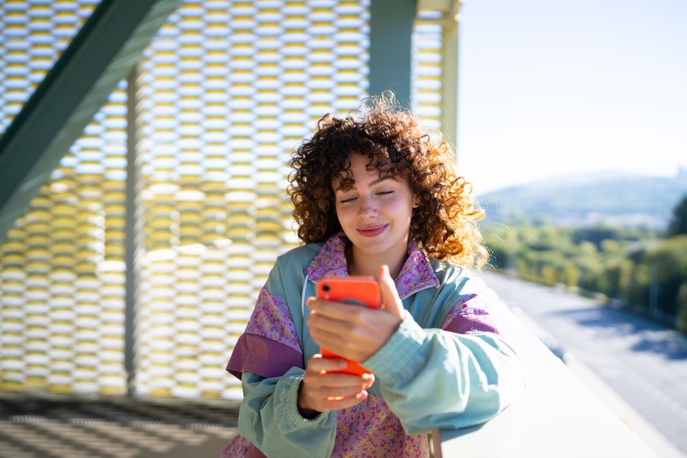 Smiling woman with curly hair using a smartphone on an outdoor balcony overlooking a scenic road in daylight, Spain.