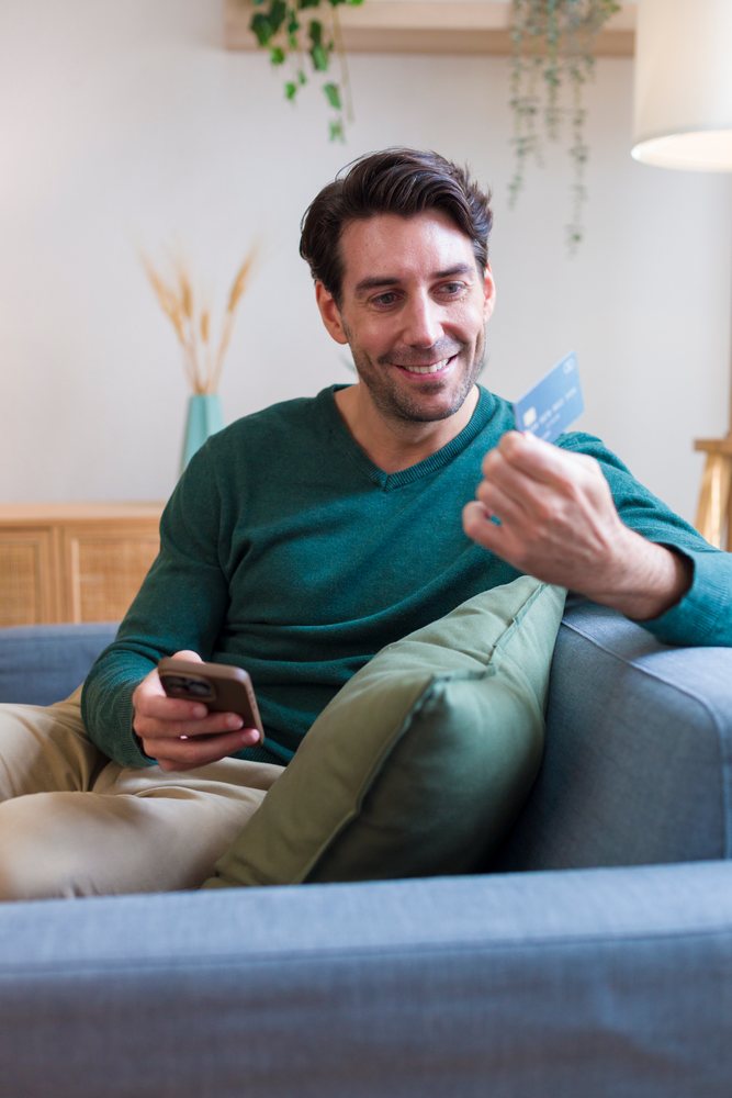 Smiling man seated on a gray couch in an indoor living room holding a credit card and mobile phone, Technology, young adult, home.