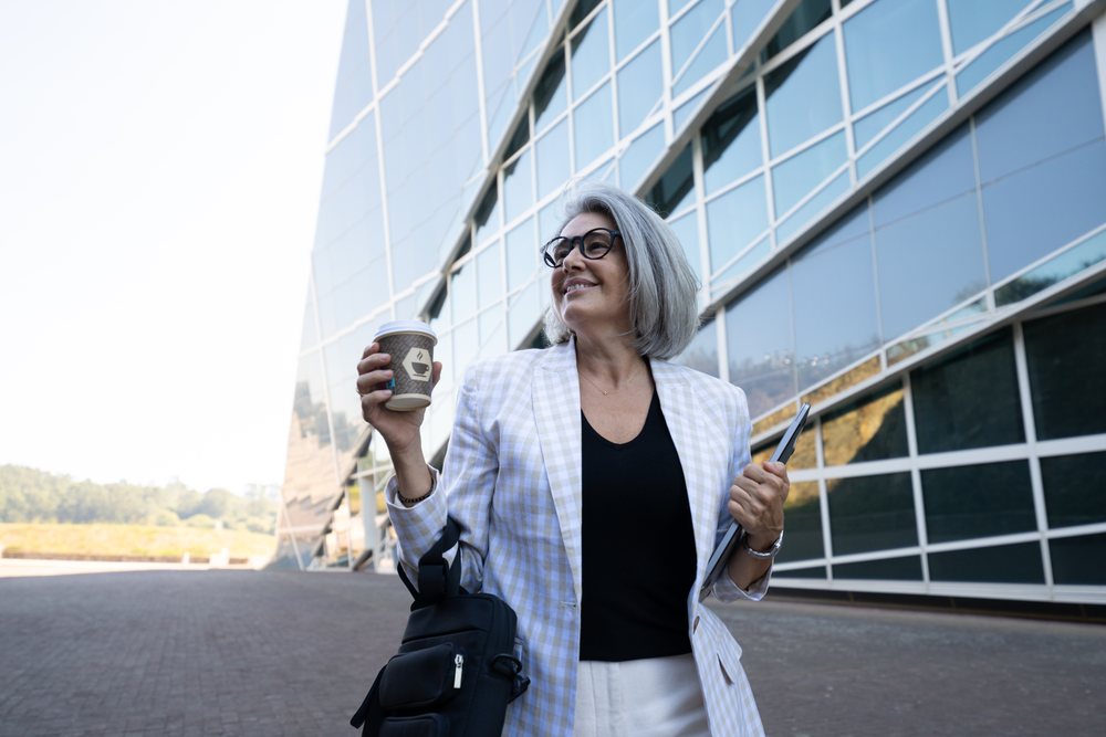 Businesswoman with gray hair smiling, holding coffee cup and tablet outside a modern office building, businesswomen, Spain.