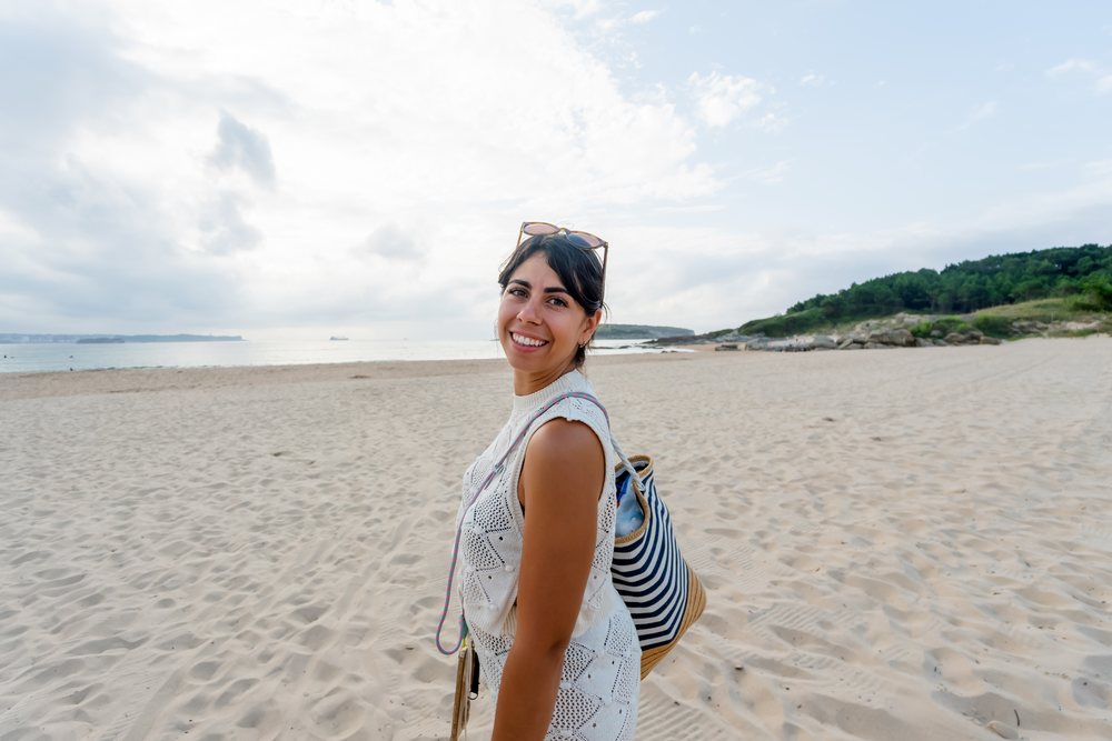 Smiling woman standing on sandy beach with sea, clouds, trees, and rocks in background during daytime, kids, Spain.