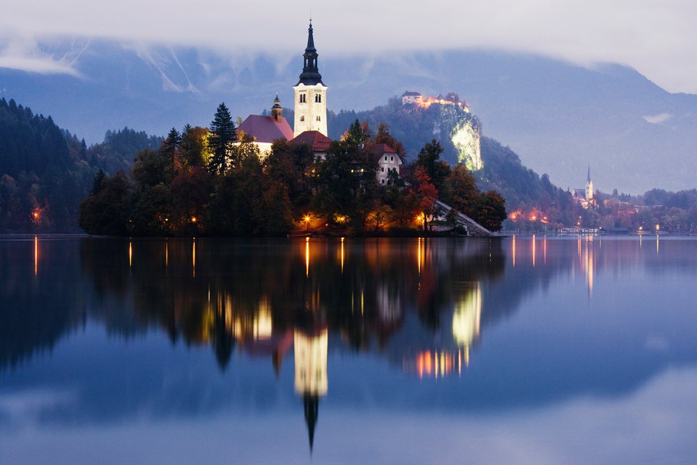 Church of Assumption illuminated on an island with mountain reflections at dusk, Lake Bled, Slovenia.