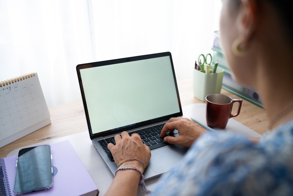 Person typing on a laptop at a desk with a coffee mug, calendar, smartphone, and stationery in a bright workspace, telecommuting, Spain.