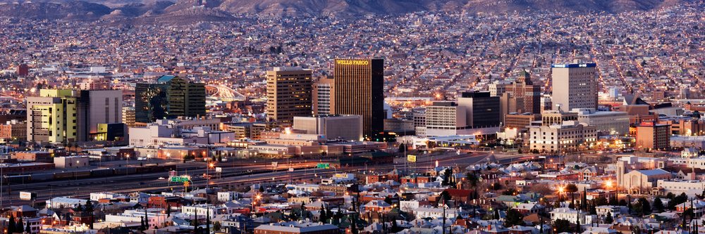 Dusk cityscape showing El Paso, Texas, and Juarez, Mexico, with illuminated buildings, urban infrastructure, and mountains in the background.