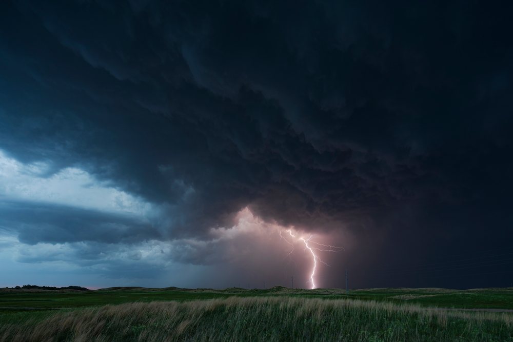 Lightning bolt strikes the ground amidst dark storm clouds over a grassy field crossed by power lines during Blue Hour, Storm Chasing, Weather, Environmental Issues, Dunning, Nebraska, USA.