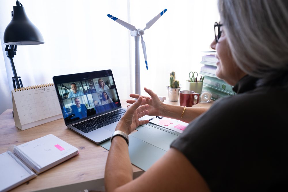 Woman wearing glasses gestures during a video call on a laptop at a desk with office supplies and a wind turbine model in a daylight-lit home office, Spain.