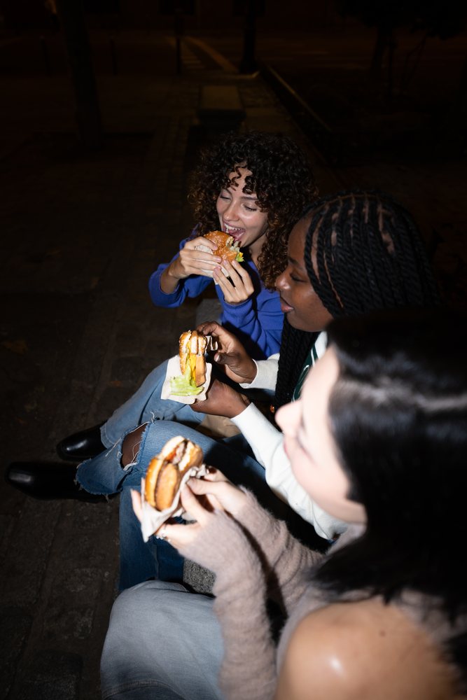 Three people sitting on a bench eating burgers outdoors at night, Madrid, Spain.