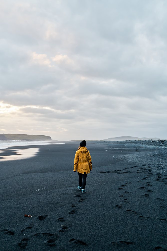 A white person walks on a black sand beach under a cloudy sky