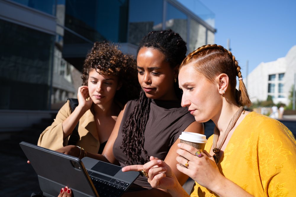 Three young women stand outdoors near a modern building, using a tablet on a sunny day, Spain.