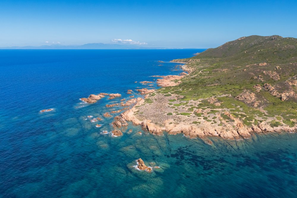 Aerial view of Cala Sarraina coastline shows rocky shore, clear blue Mediterranean Sea, and hilly terrain, Travel Images, Sardinia, Italy.