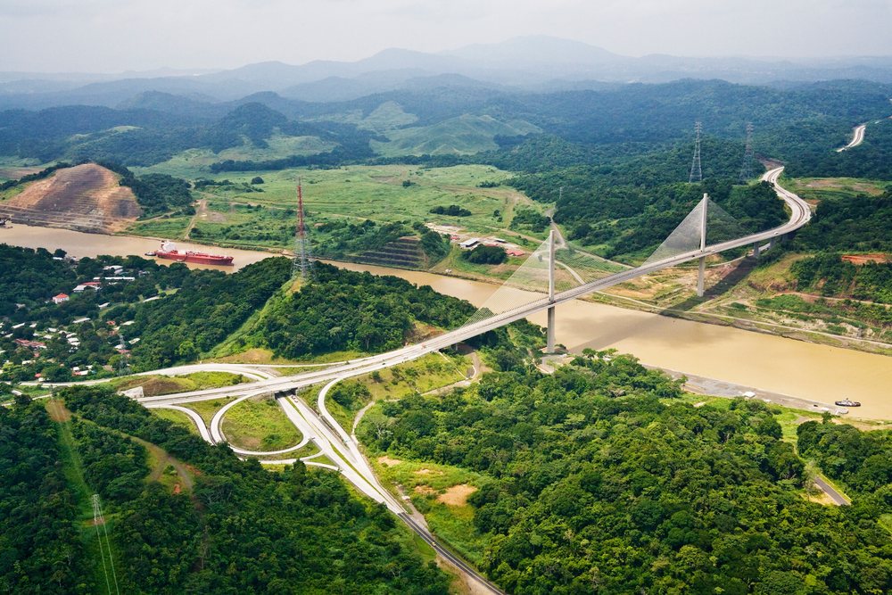 Panama Canal Bridge spans a river with a cargo ship passing beneath amid green hilly landscape and electric transmission towers.