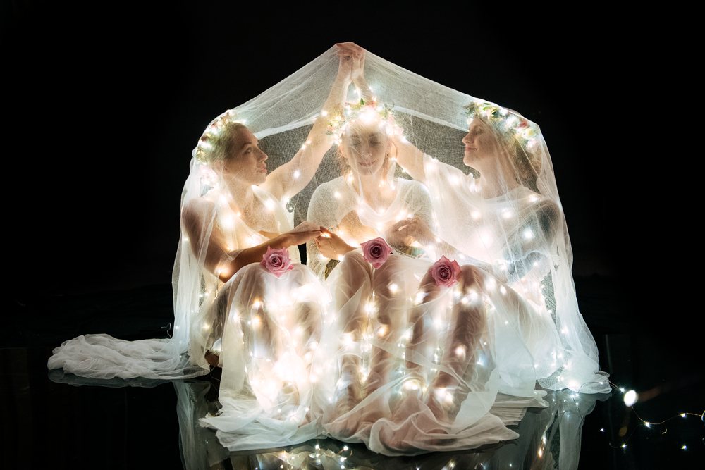 Three women wearing white dresses and floral crowns sit closely under a sheer veil wrapped with glowing fairy lights in a dark studio, USA.