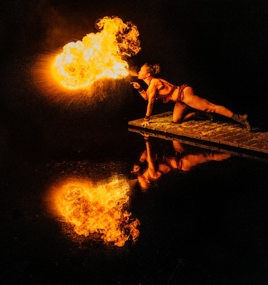 Female fire breather kneels on a platform at night, producing a vibrant flame reflected on calm water, artistry, artist, USA.