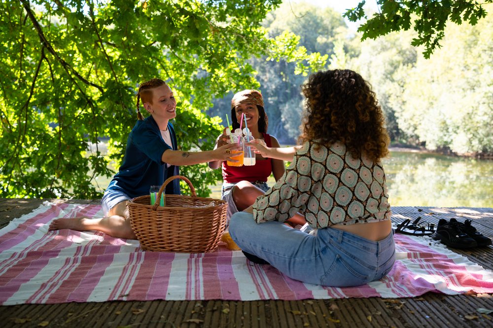Three women toast with drinks on a pink-striped blanket by a shaded riverside wooden deck in summer, Spain.
