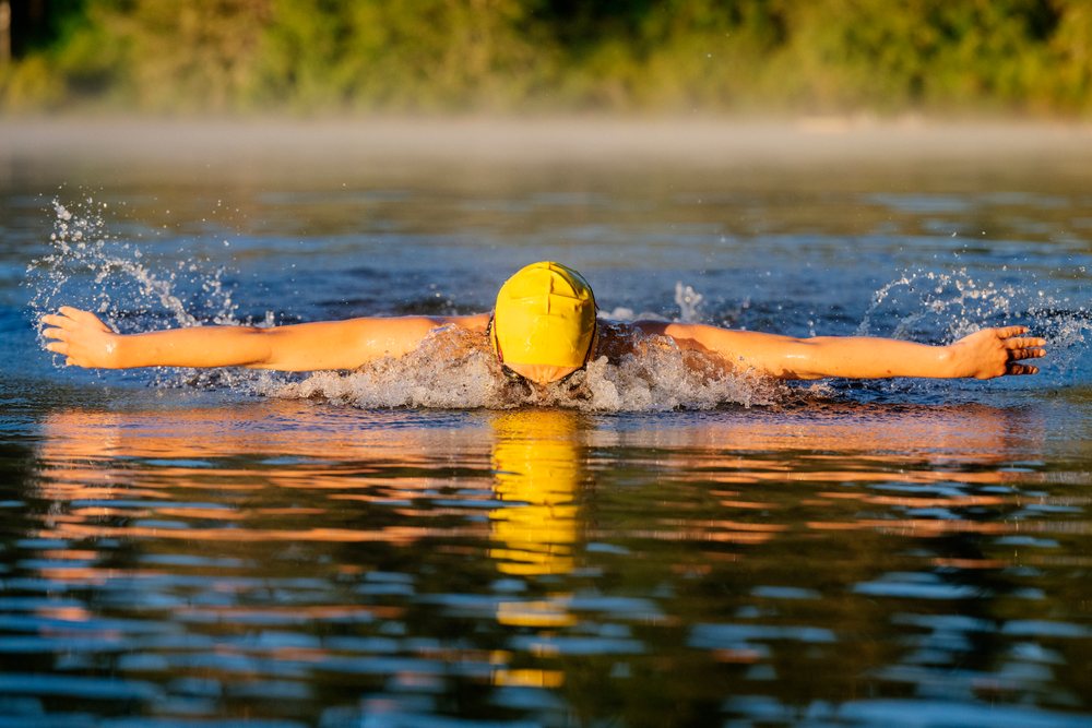 Swimmer wearing a yellow swim cap performing butterfly stroke in open water at sunrise, USA.