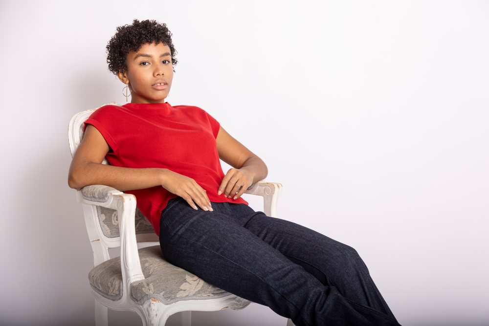 Woman wearing a red shirt and jeans sitting relaxed on an ornate upholstered chair against a white background, fashion, clothes, young culture.