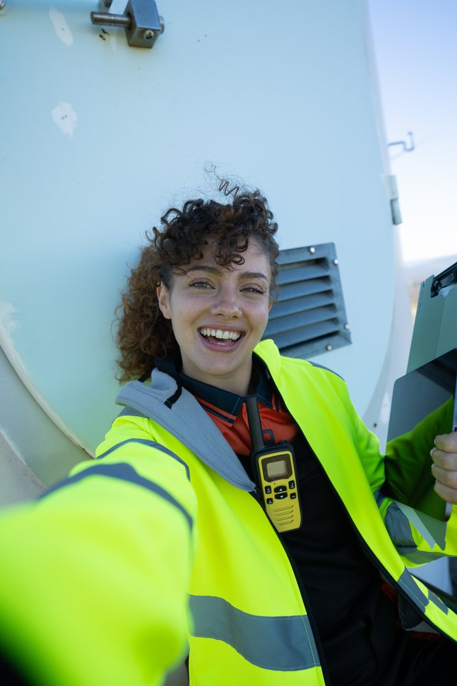 Smiling woman with curly hair wearing a high visibility jacket takes a selfie outdoors in an industrial setting while holding a clipboard and walkie talkie, sustainability, renewable energy, Galicia, Spain.