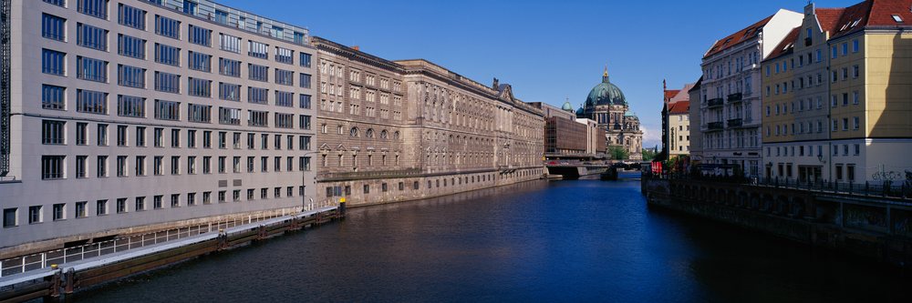 River Spree flows calmly through Berlin beneath a clear blue sky, bordered by historic and modern buildings with a distant bridge and cathedral dome, River Spree Berlin, Germany.
