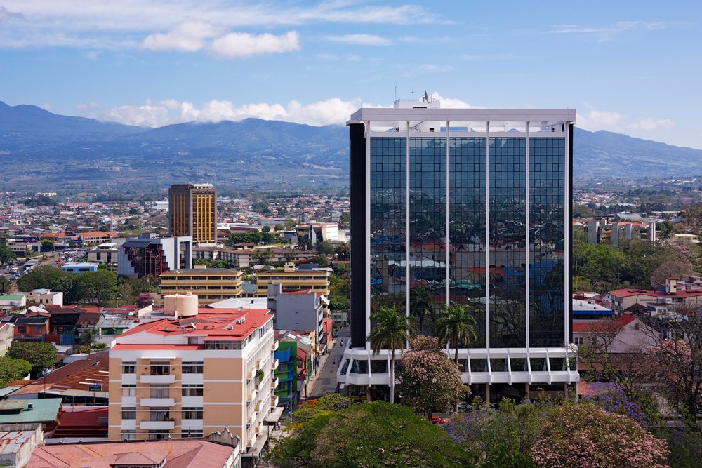 Glass facade building reflects urban cityscape with trees and mountains visible, San Jose, Costa Rica.