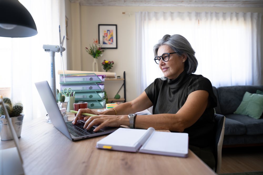 Woman with gray hair and glasses types on a laptop at a desk in a home office living room with cactus plants and folders nearby, Spain.