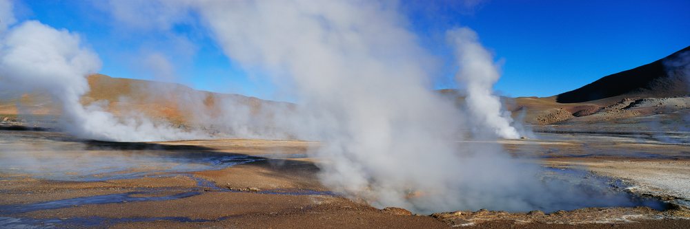 El Tatio Geysers erupting steam over arid desert terrain with mountains and clear blue sky, El Tatio Geysers Atacama Desert, Chile.