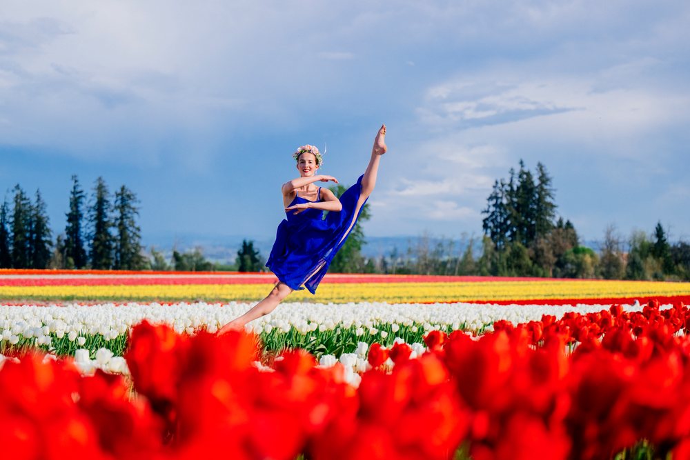 Ballet dancer in a blue dress leaps joyfully in a tulip field under a cloudy sky, USA.
