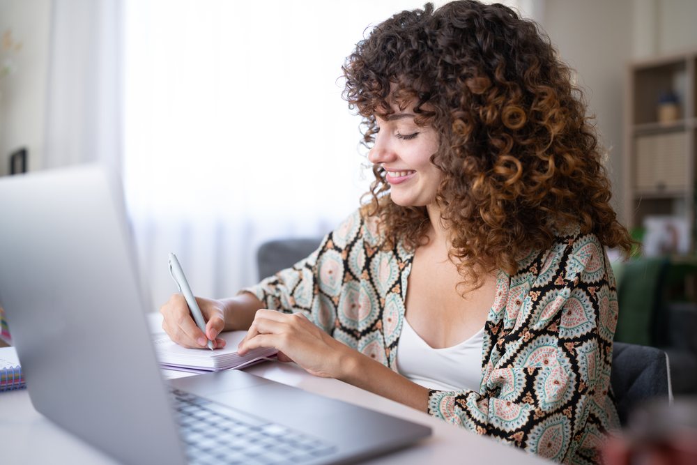 Woman with curly hair smiling and writing in a notebook at a home office desk with a laptop, Spain.