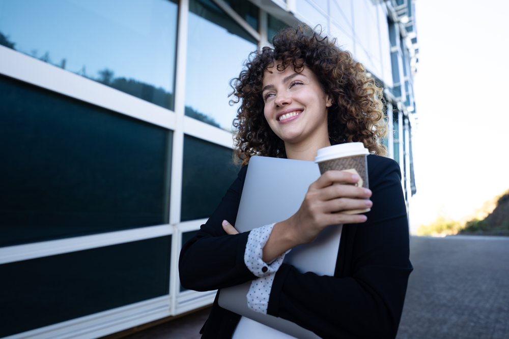 Businesswoman holding laptop and coffee cup outdoors near modern building, business trip, Spain, Santiago De Compostela.