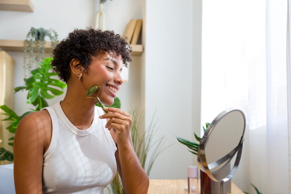 Young woman holding a facial roller near her face in front of a round mirror with green plants and natural light.