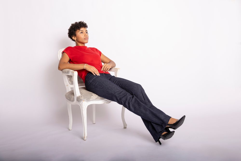 Young woman seated contemplatively on a white armchair wearing a red top, dark jeans, and black heels in a studio, fashion, clothes, young culture.