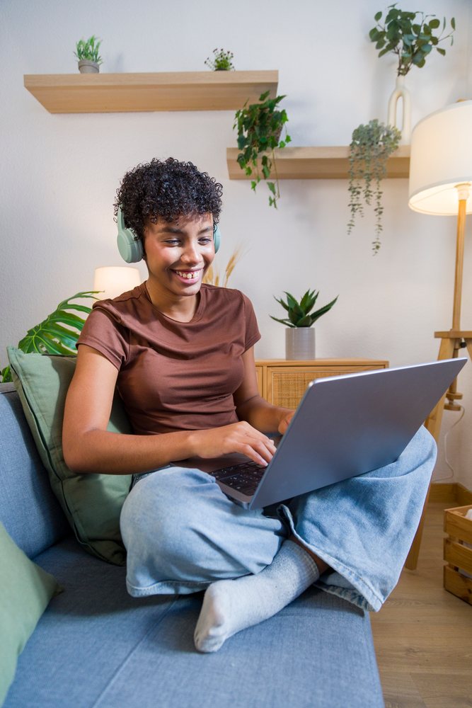 Young woman wearing headphones uses a laptop while sitting cross-legged on a sofa in a cozy living room with modern decor and indoor plants.