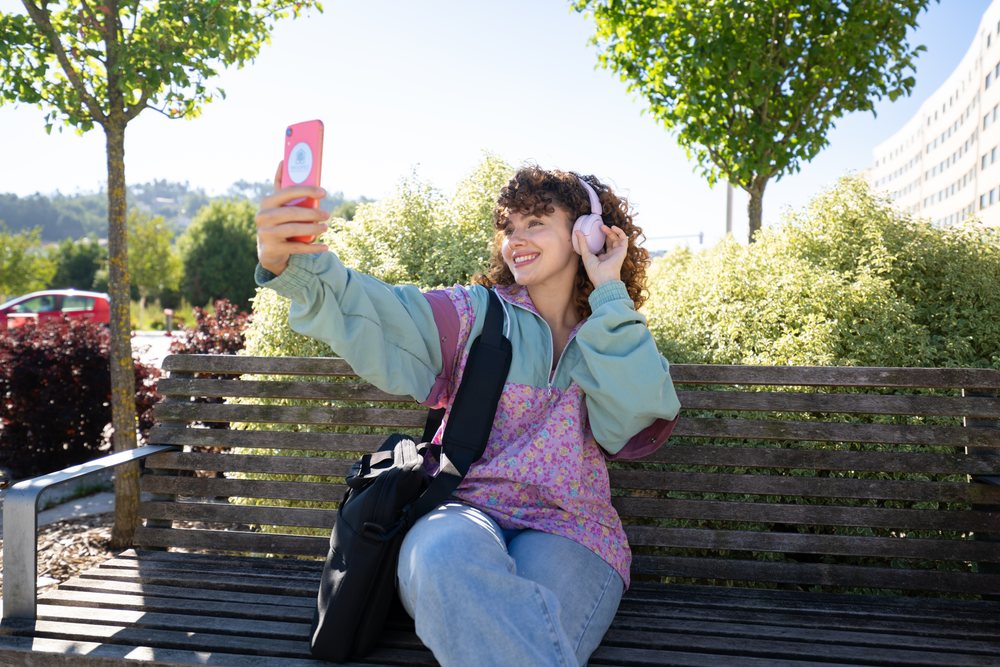 Woman wearing headphones and floral jacket taking a selfie while sitting on a wooden bench in a sunny park outdoors, Spain.
