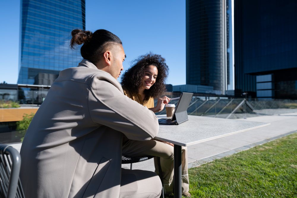 Two young adults collaborating using a tablet at an outdoor urban workspace with skyscrapers and a coffee cup on the table, Madrid, Spain.