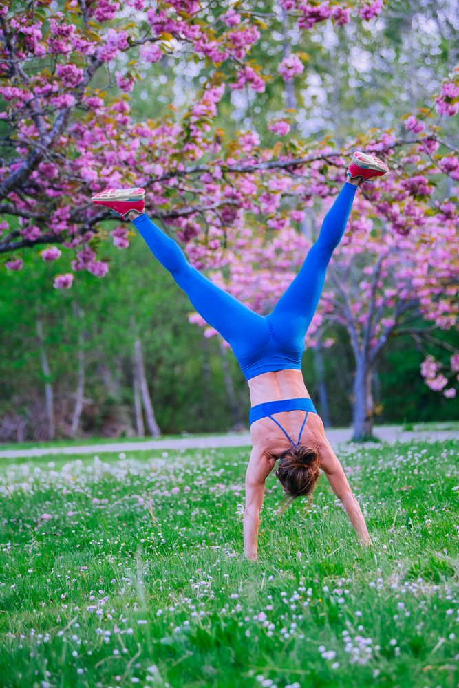 Woman in blue sportswear performs a handstand outdoors on green grass with wildflowers and pink blossoming trees in spring, Calisthenics, USA.