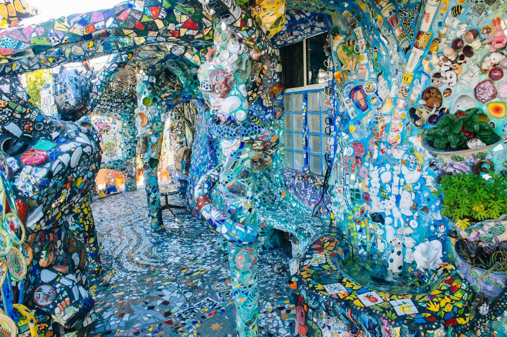 Colorful mosaic art with decorated columns, walls, floor, bench, ceramic figurines, and green plants in an outdoor space, CA, USA.
