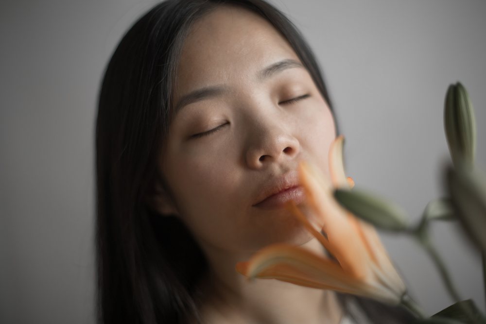 Woman with closed eyes enjoying the scent of an orange lily flower indoors with a serene expression, Art, portrait, emotional.