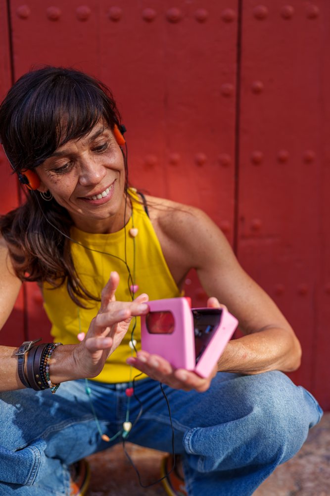 Smiling woman in a yellow tank top and blue jeans squats outdoors holding a pink instant camera against a red wall, retro, vintage, Málaga, Spain.