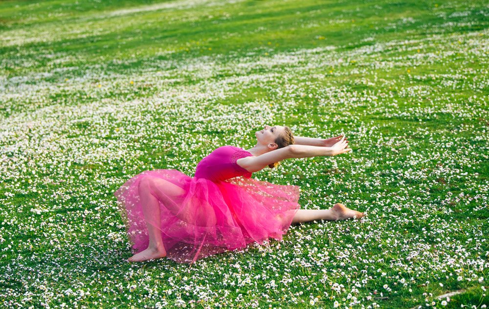 Barefoot woman wearing a pink tutu stretches in an outdoor green field dotted with white daisies under daylight, USA.