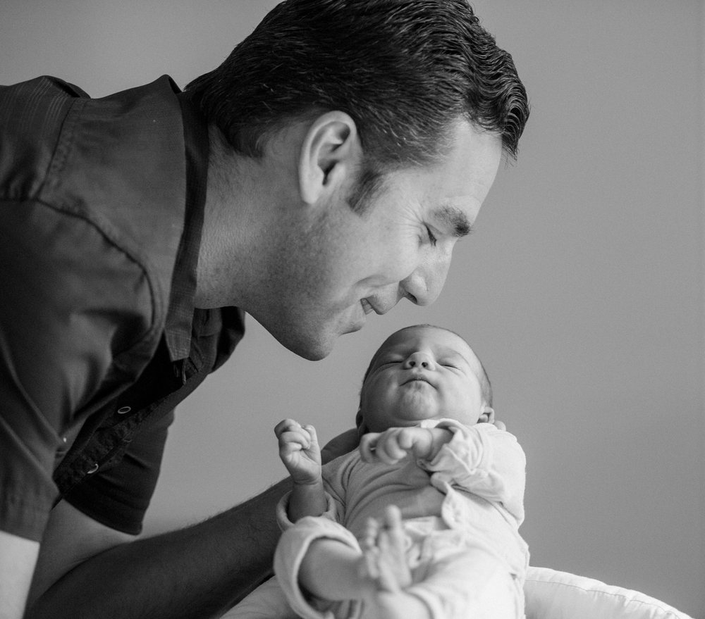 Father lovingly smiles while closely holding his sleeping newborn baby indoors, USA.