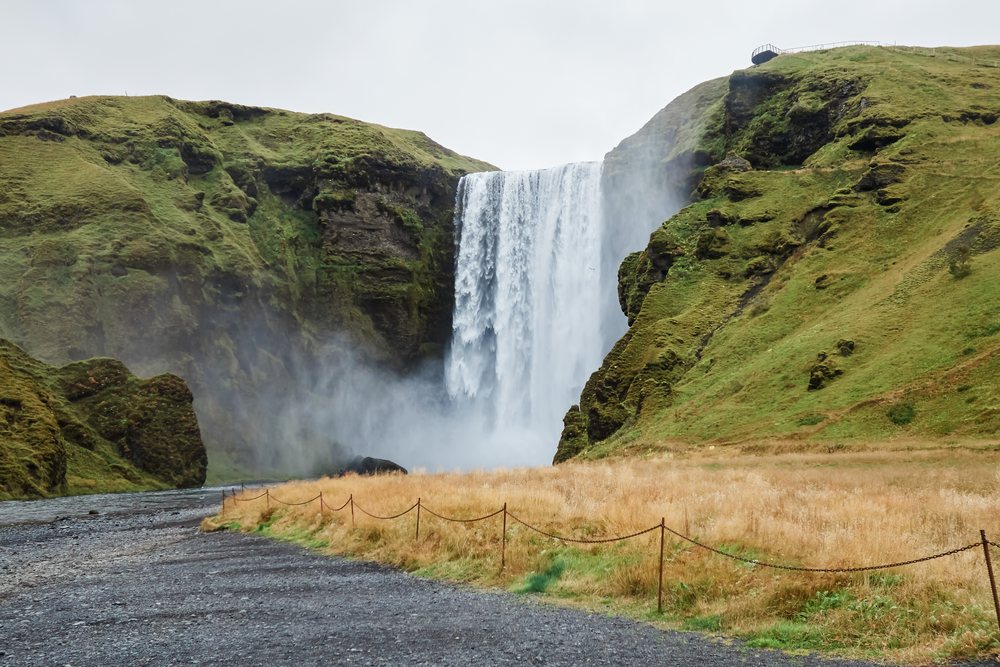 Waterfall cascades down a moss-covered cliff surrounded by grassy hills and a stone path, Iceland.