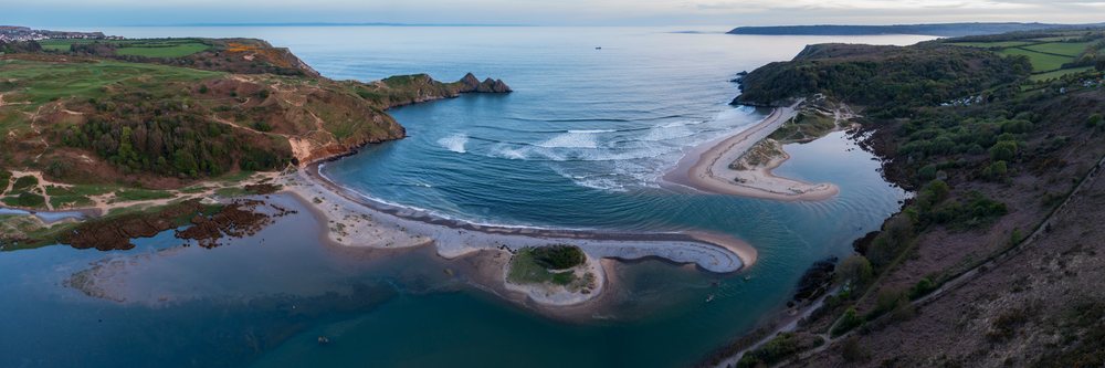 Aerial view of three Cliffs Bay showing sandy beach, waves, rocky cliffs, and grassy hills on the Gower Peninsula, Landscape, Swansea, Glamorgan, Wales.