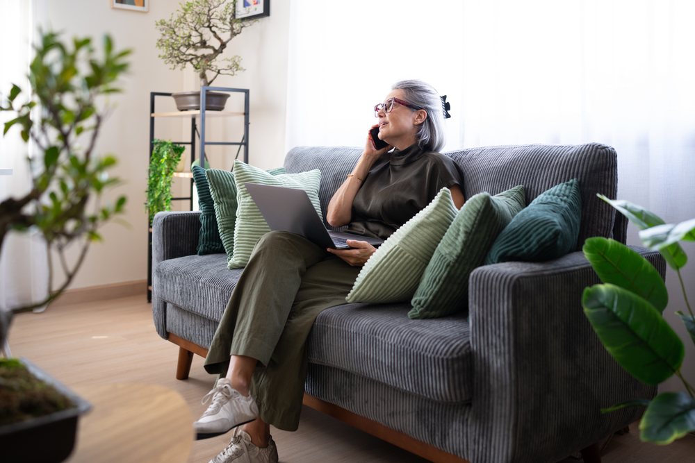 Senior woman seated on a gray sofa talking on the phone and using a laptop in a cozy indoor living room with plants, Spain.