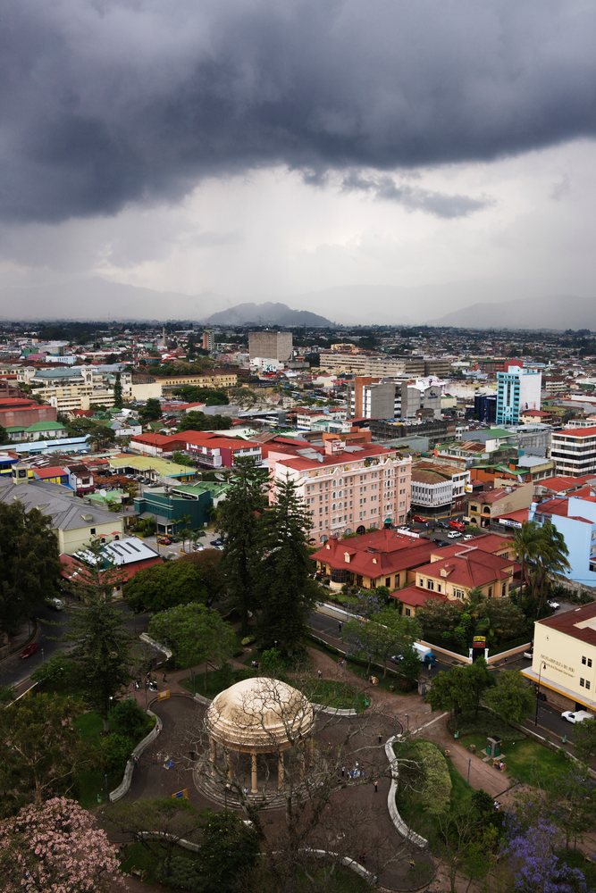 Storm clouds gather above San Jose city showing an urban park with a dome gazebo, various buildings, roadways with vehicles, and mountains in the distance, Costa Rica.