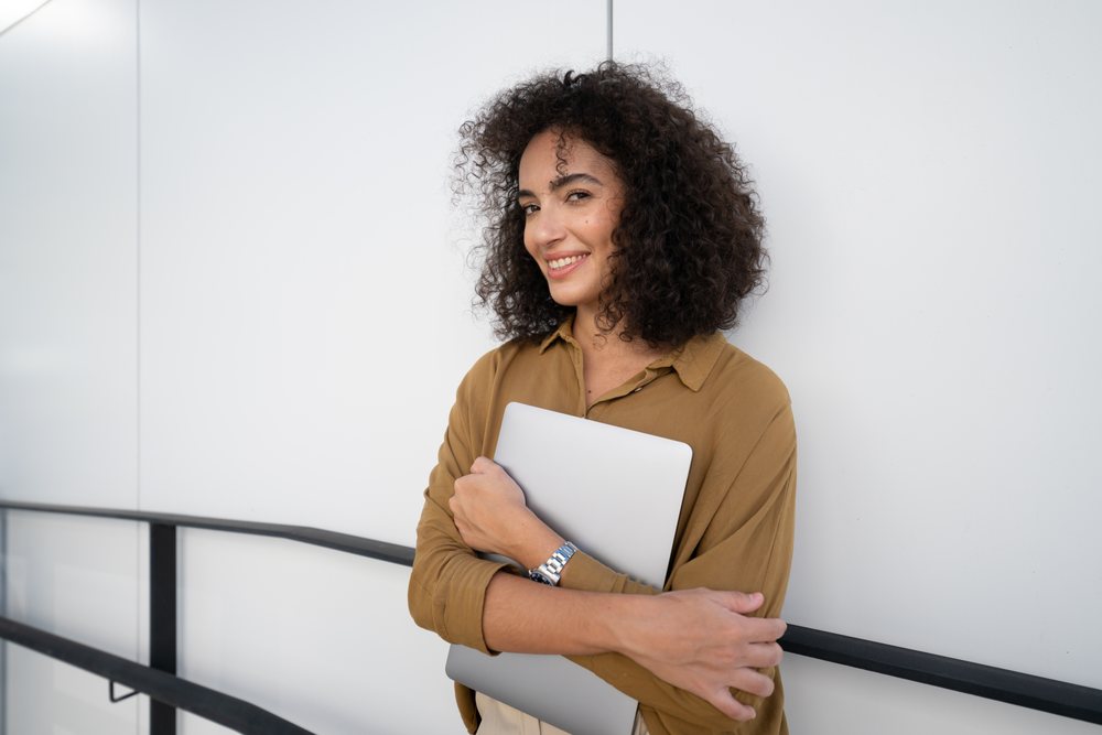 Woman with curly hair and a brown long-sleeve shirt smiles while holding a laptop and leaning against a black railing near a white wall outdoors, Madrid, Spain.