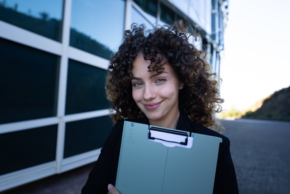 Businesswoman holding a clipboard and smiling outdoors near a modern building during a business trip, Spain, Santiago De Compostela.