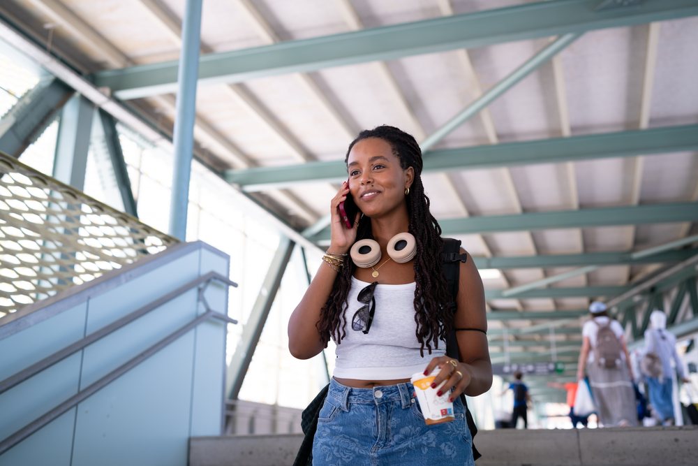 A young woman wearing a backpack and headphones talks on her phone while holding a drink in a bus station, remote work, travel, Santiago De Compostela, Spain.